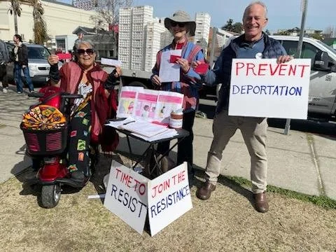 RAEB volunteers at a farmers market with Prevent Deportation and Join the Resistance signs